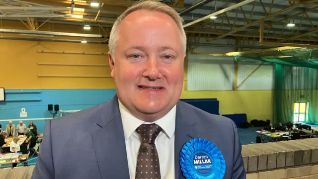 Darren Millar is wearing a suit and a blue lapel, he's standing in a sports hall.