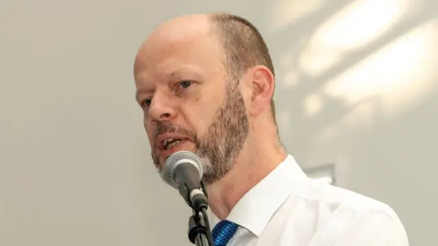 Jamie Driscoll, who is wearing a white shirt and a blue tie, is speaking into a microphone. He is balding with very short cropped brown hair at the sides and a beard.