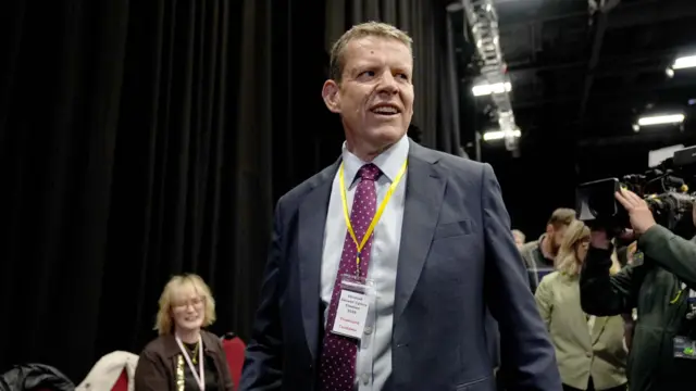Plaid Cymru leader Rhun ap Iorwerth arrives at the Bangor Conwy Mon election count. He is wearing a grey suit and a red tie.
