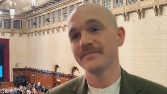 Luke Reynolds stood in a hall with people in the background