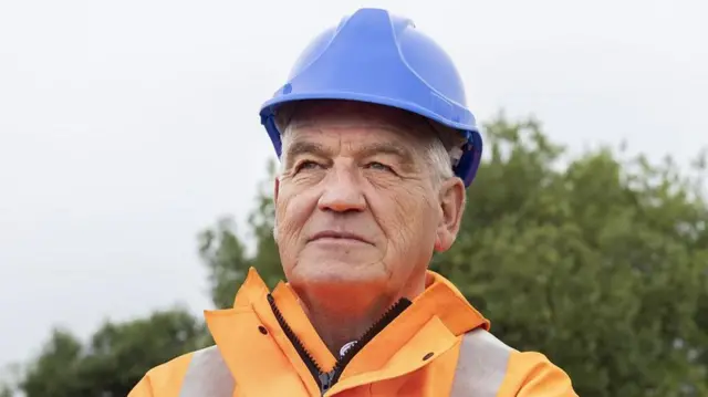 Gateshead Council leader Martin Gannon at the Leamside Line, wearing a blue hard hat and orange hi-vis jacket. He is staring into the distance.
