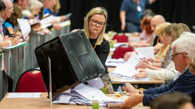 A woman tips out a container of ballots onto a table of people counting