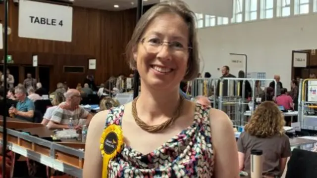 A woman in a floral vest and brown bead necklace with a yellow rosette stands in a hall with people counting at tables behind her. She has short brown hair and wireless glasses.