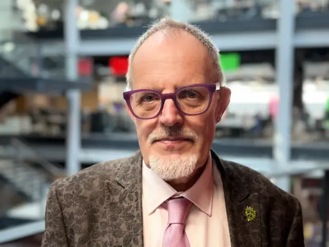 Ian Chandler wears purple glasses and a flowery grey suit with a pink tie. He has short white hair and a short white beard.