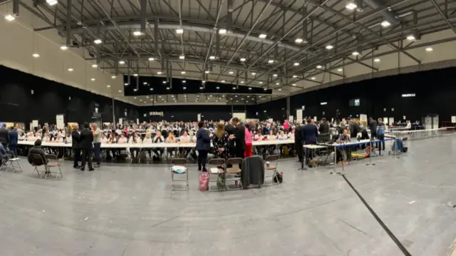 An large hall concourse, with people sitting at long table and others standing.