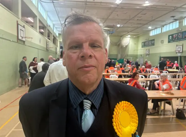 Lib Dem Julian Cooper posing for a picture after the vote count. There are many people sitting behind him.