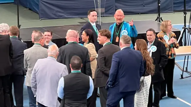 A group of people stand in front of the stage in the sports hall. At least four of them are wearing turquoise rosettes for Reform.