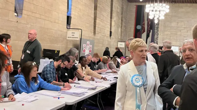 An election count. There are dozens of people sitting in a row on one side of a white table, which is stacked with ballot papers. A sign above them reads: "Dene and South Gosforth". There are groups of people standing on the other side of the table, including a woman in a white suit and a large, turquoise rosette for reform.
