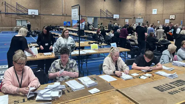 There are rows of people sitting at wooden tables snaking around the red-bricked sports hall. There are counting ballot papers in front of them. There are various people standing around too, observing.