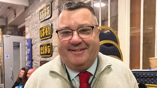 Kevin Small smiles at the camera - he wears glasses, a beige fleece and Labour rosette.