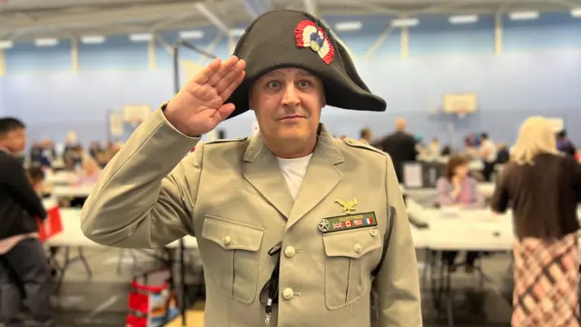 Pascual Jean Louis wearing a French style military uniform and saluting, while stood in a leisure centre hall.