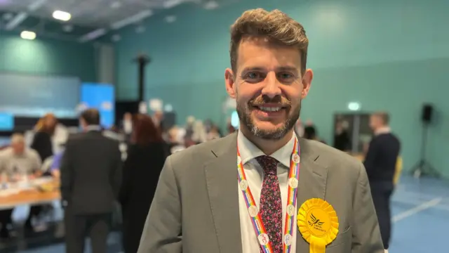 A man in a grey suit with a rainbow lanyard and a yellow rosette smiles at the camera