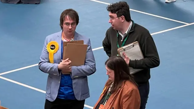 Paul Edgeworth is watching the counting while speaking with someone on his right. He is wearing a yellow rosette for the Liberal Democrats and a light blue suit.