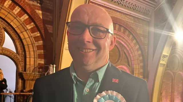 A man smiles at the camera. He is bald and wears glasses, a suit jacket and green shirt.