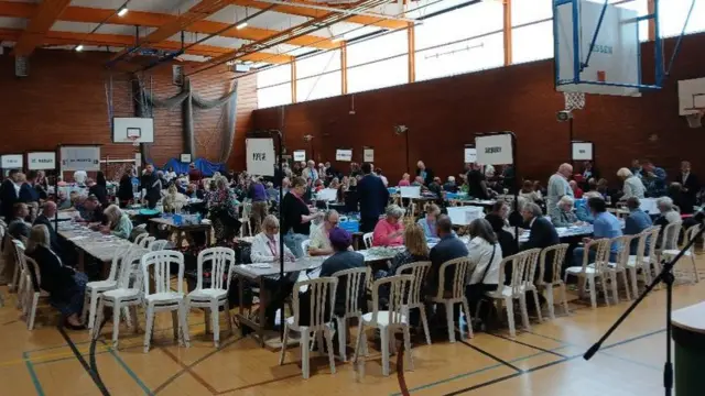 People sit at long tables inside a sports hall, with flags marking particular areas.