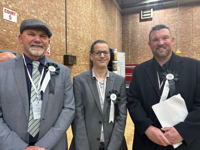 Three men, wearing black rosettes, smile into the camera