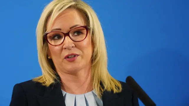 First Minister Michelle O'Neill speaking at a news conference, against a bright blue background