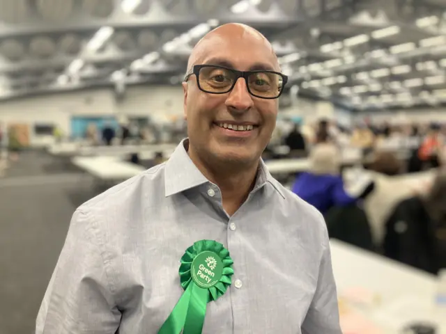 Nilesh Chohan, a South Asian male wearing a grey shirt and green rosette.