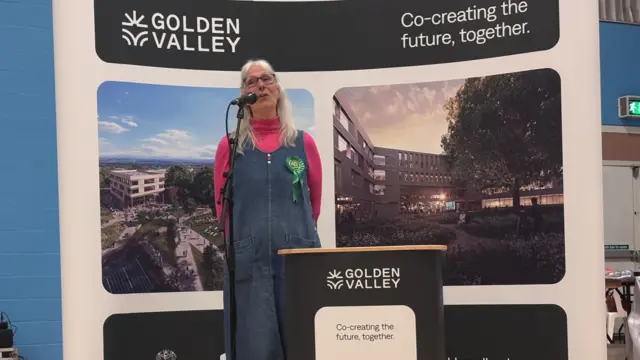 Jan Foster standing behind a podium wearing a denim dress, pink turtle neck jumper and a Greens party rossette.