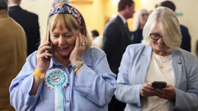 A woman wearing a Reform UK rosette speaks on the phone while another woman behind her looks at her mobile