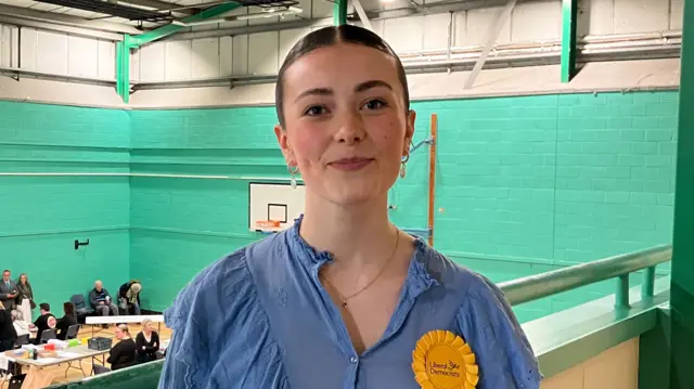 A young woman smiles at the camera as she stands on a balcony above a sports hall. She has dark hair that has been tied back and wears a blue blouse with a yellow rosette.