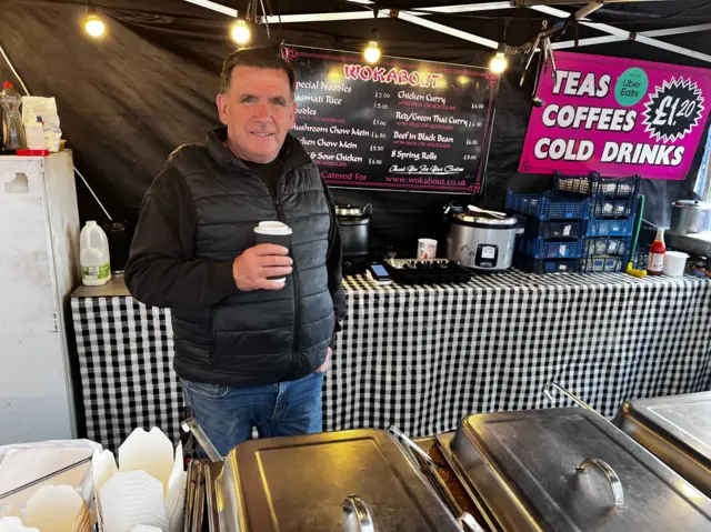 A man standing beside a food stall he is wearing a black gilet and is holding a disposable coffee cup.