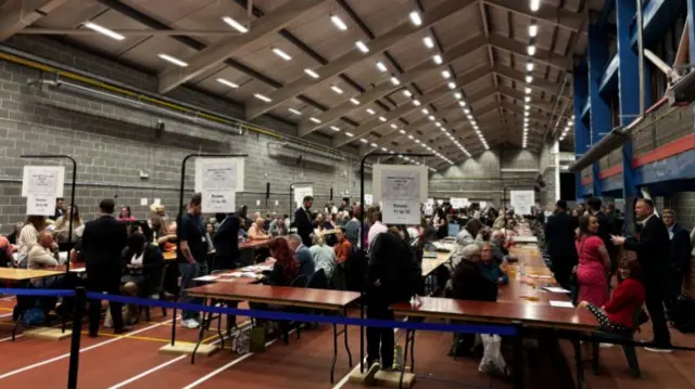 A sports hall filled with tables and people counting ballots at them