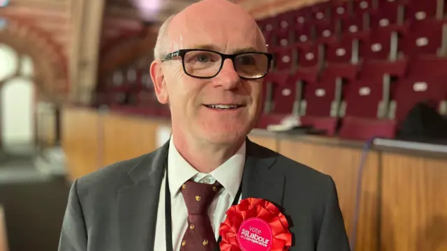 Neil MacDonald looks to the right on the camera and smiles. He is mainly bald and wears glasses with a suit and red rosette.