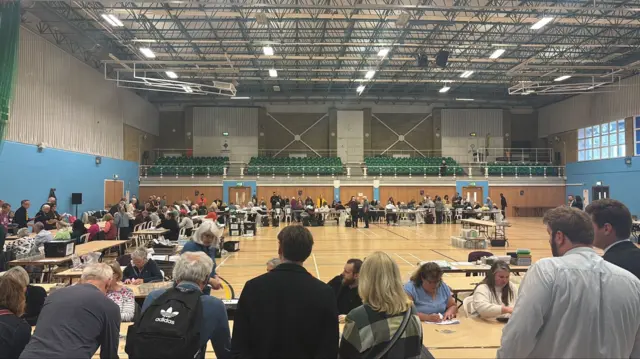 The verification count in Cheltenham. They are in a sports hall, with people sat at desks.