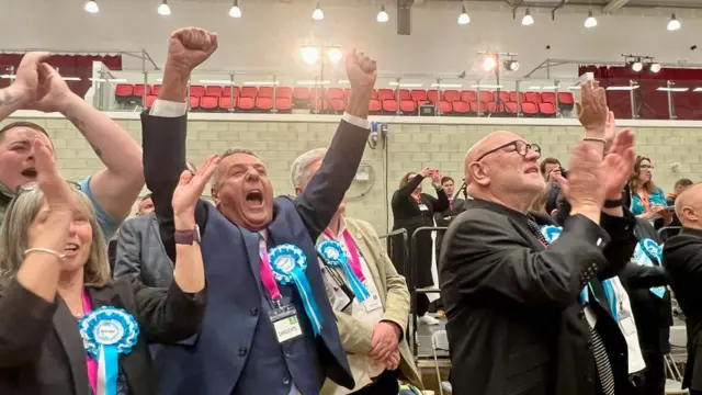 A number of campaigners wearing Reform rossettes celebrate and cheer in a town auditorium