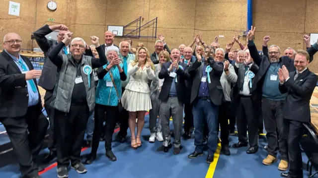 Wide shot of a large group of people standing together inside a sports hall, with high brick walls and red metal roof beams overhead. The group is arranged in a semicircle on a blue floor marked with court lines, surrounded by long tables and chairs. Many people are wearing jackets and badges or rosettes, with several raising their arms or clapping. Additional equipment, signage, and a clock are visible on the walls in the background.