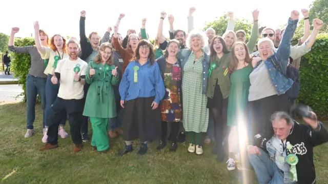 A group of male and female supporters of the Green party punch the air in celebration at their election success, as they stand on grass with a large bush seen behind them