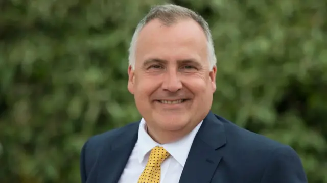 Mark Williams smiles at the camera. He has short grey hair and wears a navy suit with a yellow tie.