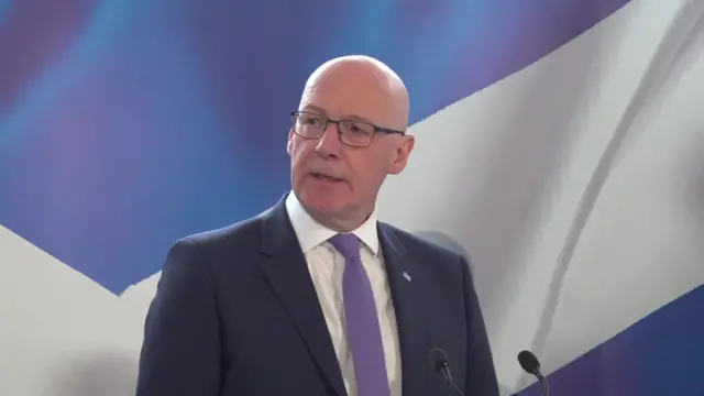 Scottish First Minister John Swinney delivers speech from a stage, two thin black mics in front of him. He's wearing a dark blue suit, white shirt and lavender tie, a Scottish Saltire (blue background with white diagonal cross) painted on a wall behind him