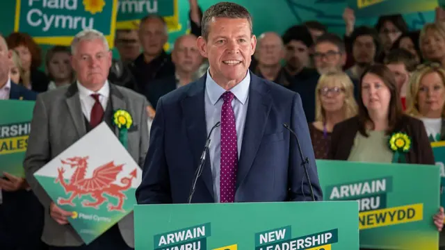 Rhun ap Iorweth in front of his party. He is wearing a blue suit and red tie, and is standing in front of a green banner that says 'Arwain Cymru O'r Newydd/ New leadership for Wales'.