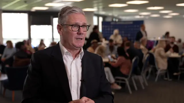 Sir Keir Starmer standing in a large room being interviewed. He is wearing a dark suit with a white shirt. There are people seated at tables in the background.