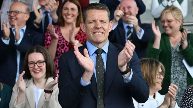 Rhun ap Iorwerth claps in the foreground, as the other Plaid Cymru MSs clap behind him all grinning.
