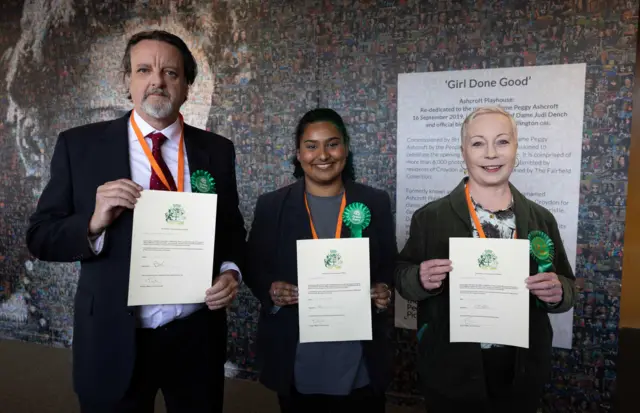 Three Green councillors with green rosettes hold up their declaration papers at a count in Croydon