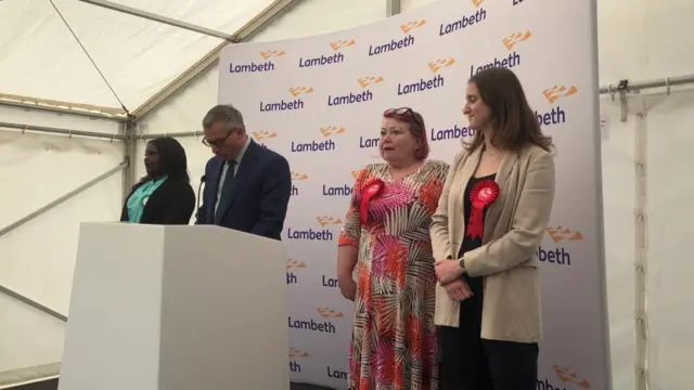 Three women stand on stage beside a podium where a man is reading out an election result. They are in a white marquee.