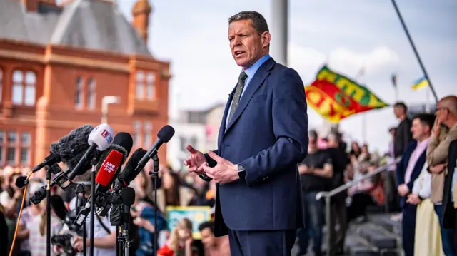 A middle aged man, wearng a navy suit, blue shirt and black and white polka dot tie. He is standing outside the Senedd and speaking into a cluster of microphones. People are gathered both in front and behind him.The red-brick Pierhead building is visible in the background.