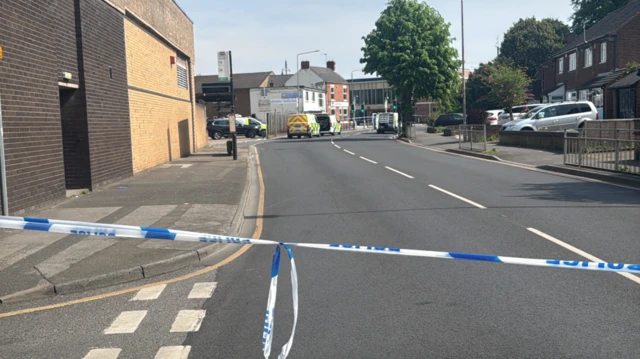 Police tape across a road with police vans visible in the background