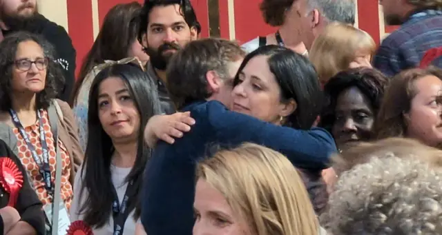 A man and a woman hug surrounded by other people at an election count inside a municipal building