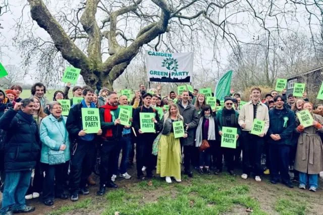 A group of Green Party campaigners pose for a photo in a park. They are holding placards and flags.