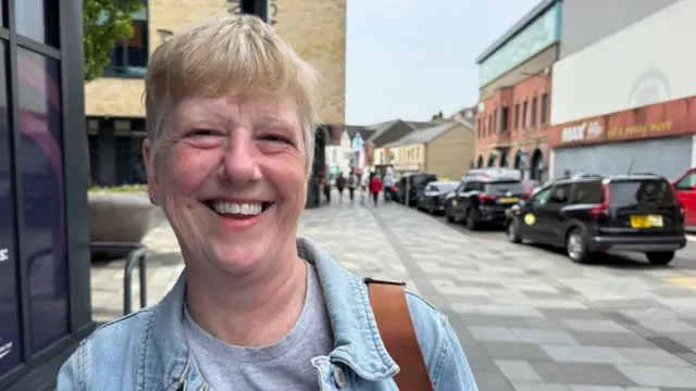 A woman with short fair hair wearing a grey t shirt and light blue denim jacket with a colourful bucket hat shaped pin badge on it. She is smiling and looking at the camera. She is stood on a street with buildings and parked cars behind her.