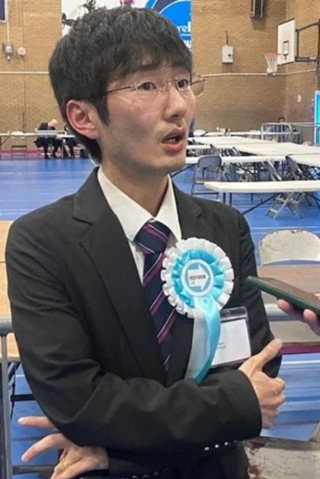 Joshua Kim wears a dark suit, a Reform rosette in a sports hall