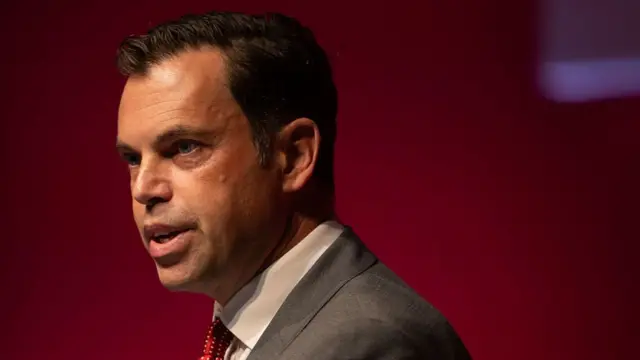 Ken Skates looks to the left, he has dark brown hair and is wearing a grey suit jacket and a red tie.