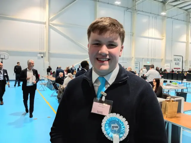 A smiling young man wearing a dark jumper with a white tie and a Reform rosette smiles for the camera as he poses inside an election count.