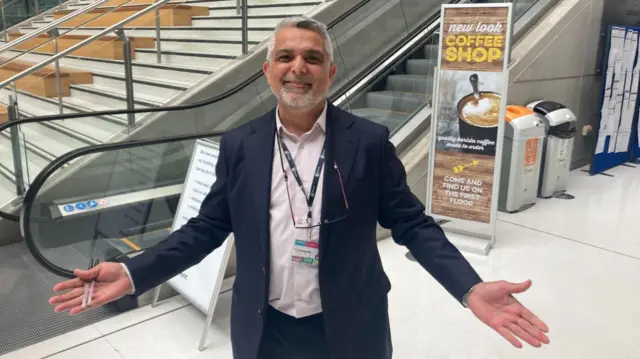 A middle aged man with a grey beard wearing a dark suit with a white shirt is holding his hands out and smiling. There is an escalator in the background.