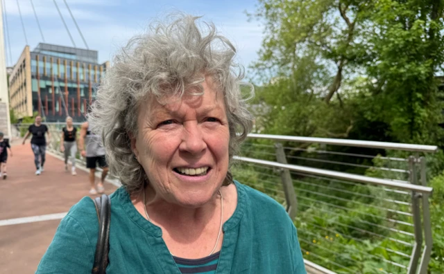 An older woman with curly grey hair, wearing a turquoise shirt. She is looking at the camera and stood on a footbridge with greenery on one side of her and other people walking behind her.