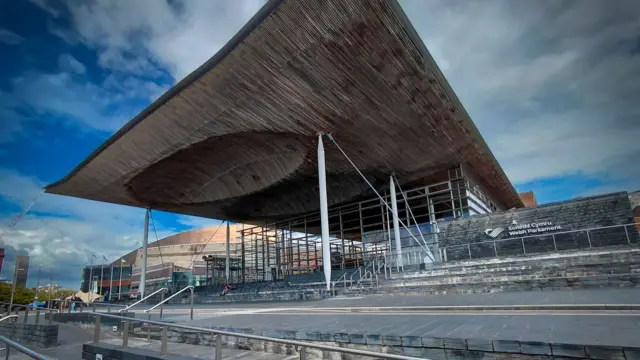 A wide‑angle exterior view of the Senedd Cymru, the Welsh Parliament building in Cardiff Bay. The image is taken from a low angle, emphasizing the large, curved wooden roof that projects outward like a canopy and is held up by slender white columns and steel cables. Beneath the roof is a glass‑fronted entrance with visible structural beams. Wide grey stone steps and gently sloping ramps fill the foreground, leading up to the building and suggesting step‑free access. The words “Senedd Cymru / Welsh Parliament” are visible on the stone wall to the right of the entrance. The sky above is dramatic, with dark grey clouds mixed with patches of blue, creating a striking contrast with the warm wood tones and cool stone surfaces of the building.